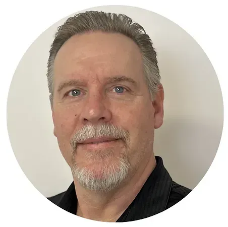 Headshot of a man with short gray hair and a goatee, wearing a black shirt against a plain light background