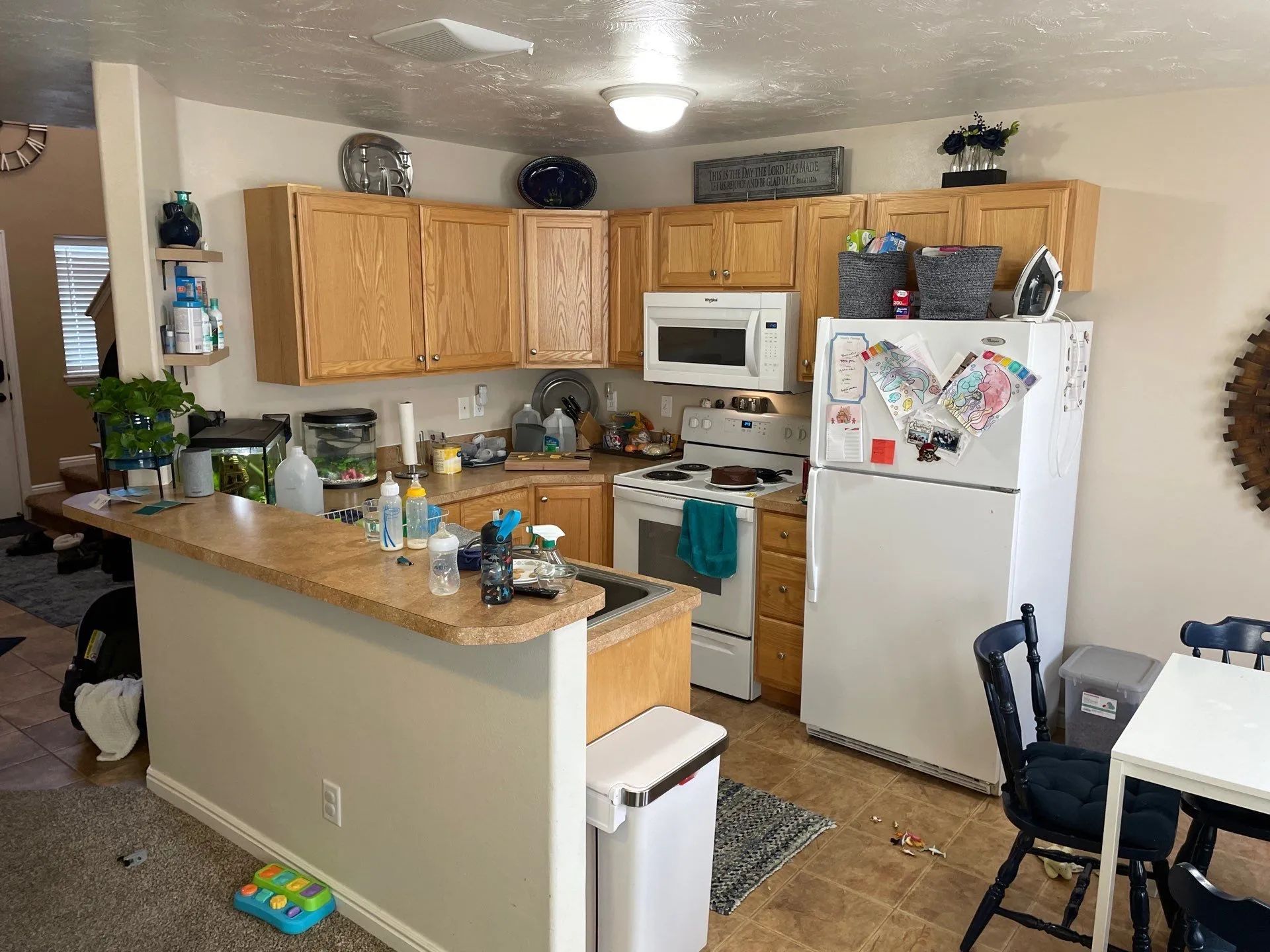 Small kitchen with wooden cabinets, white appliances, and a cluttered counter with dishes and plants.