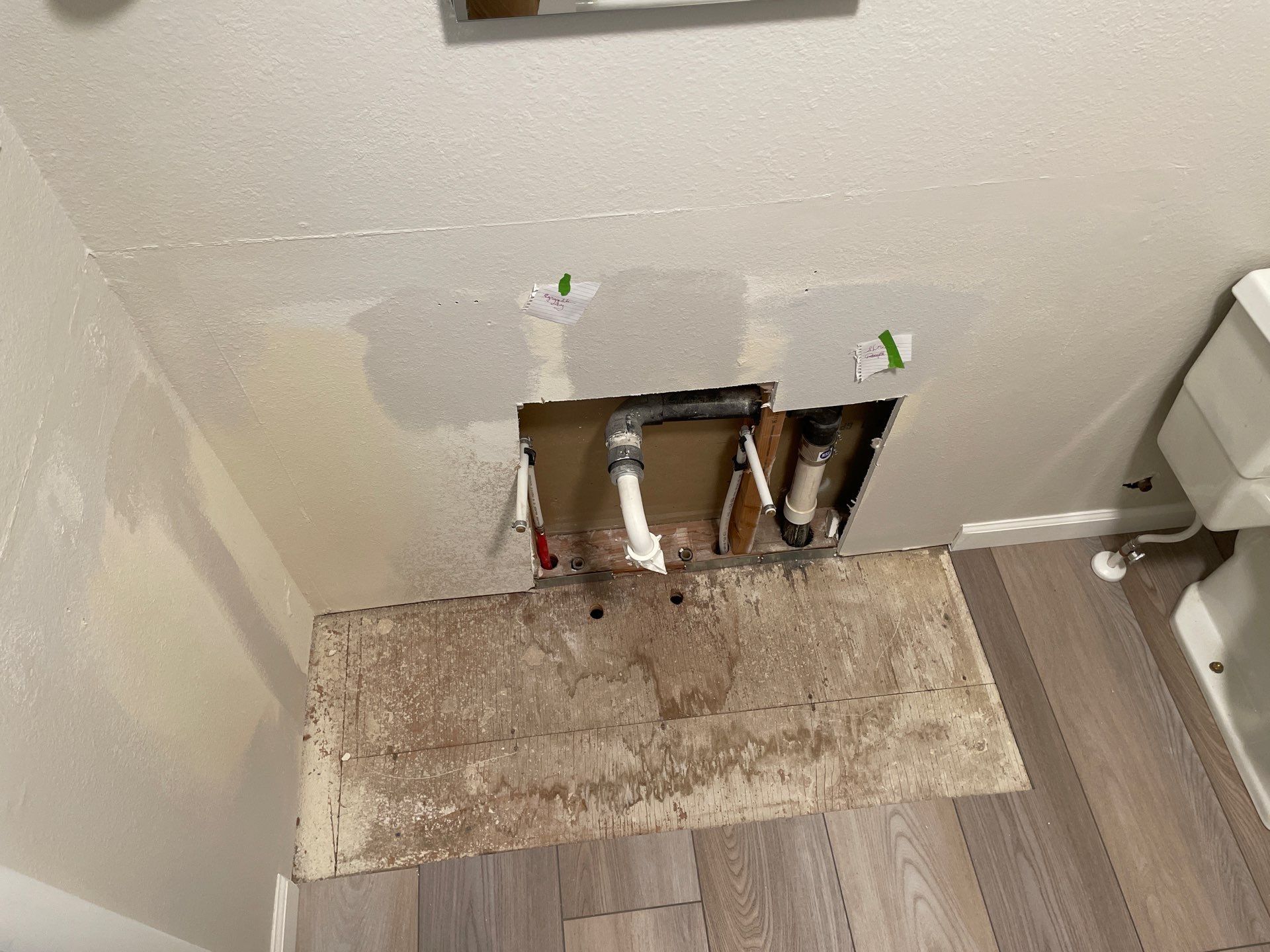 Damaged wall with exposed plumbing pipes and a missing baseboard section above a dusty floor.
