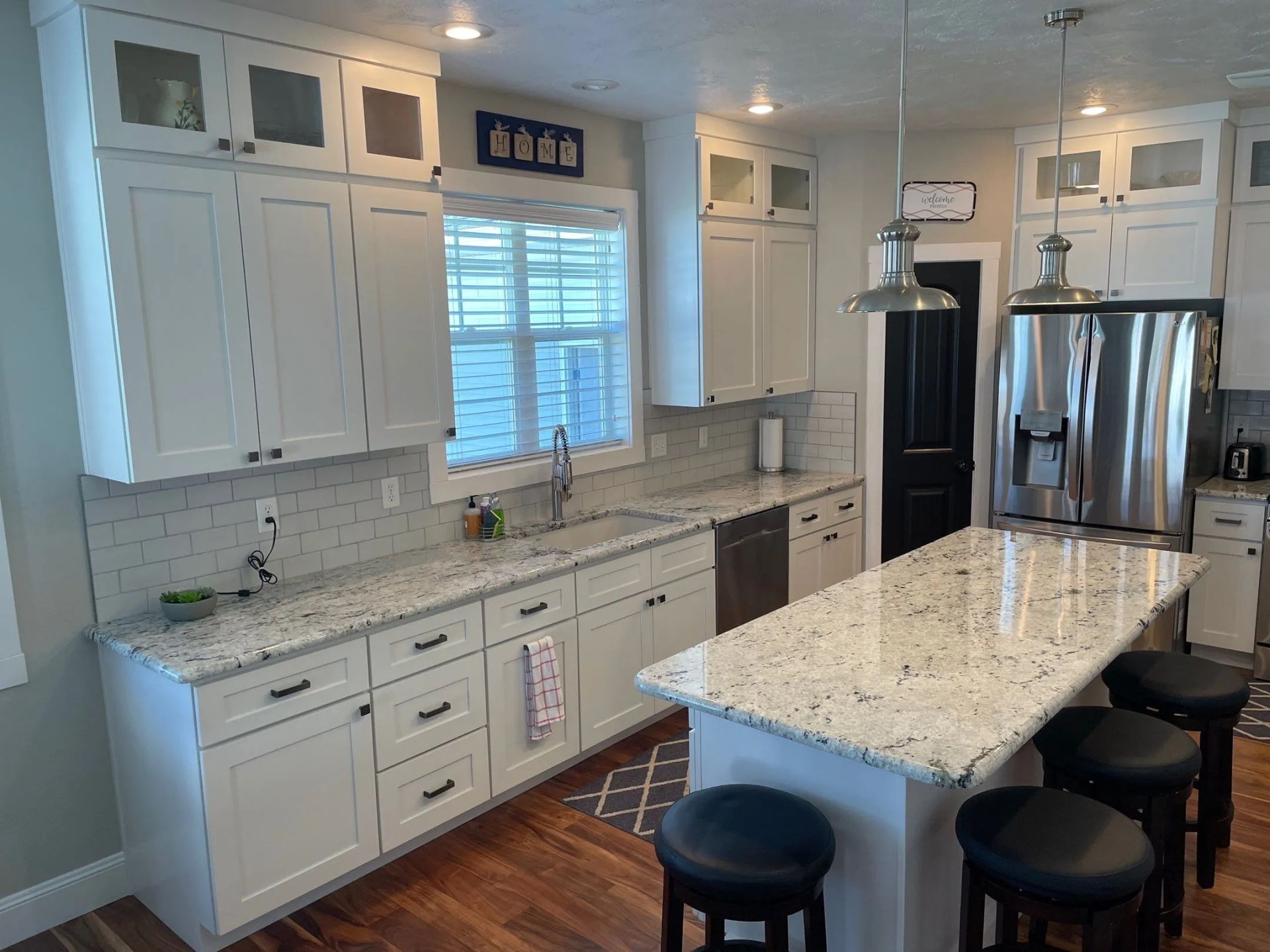 Bright white kitchen with granite island, stainless steel fridge, and black stools by the counter.
