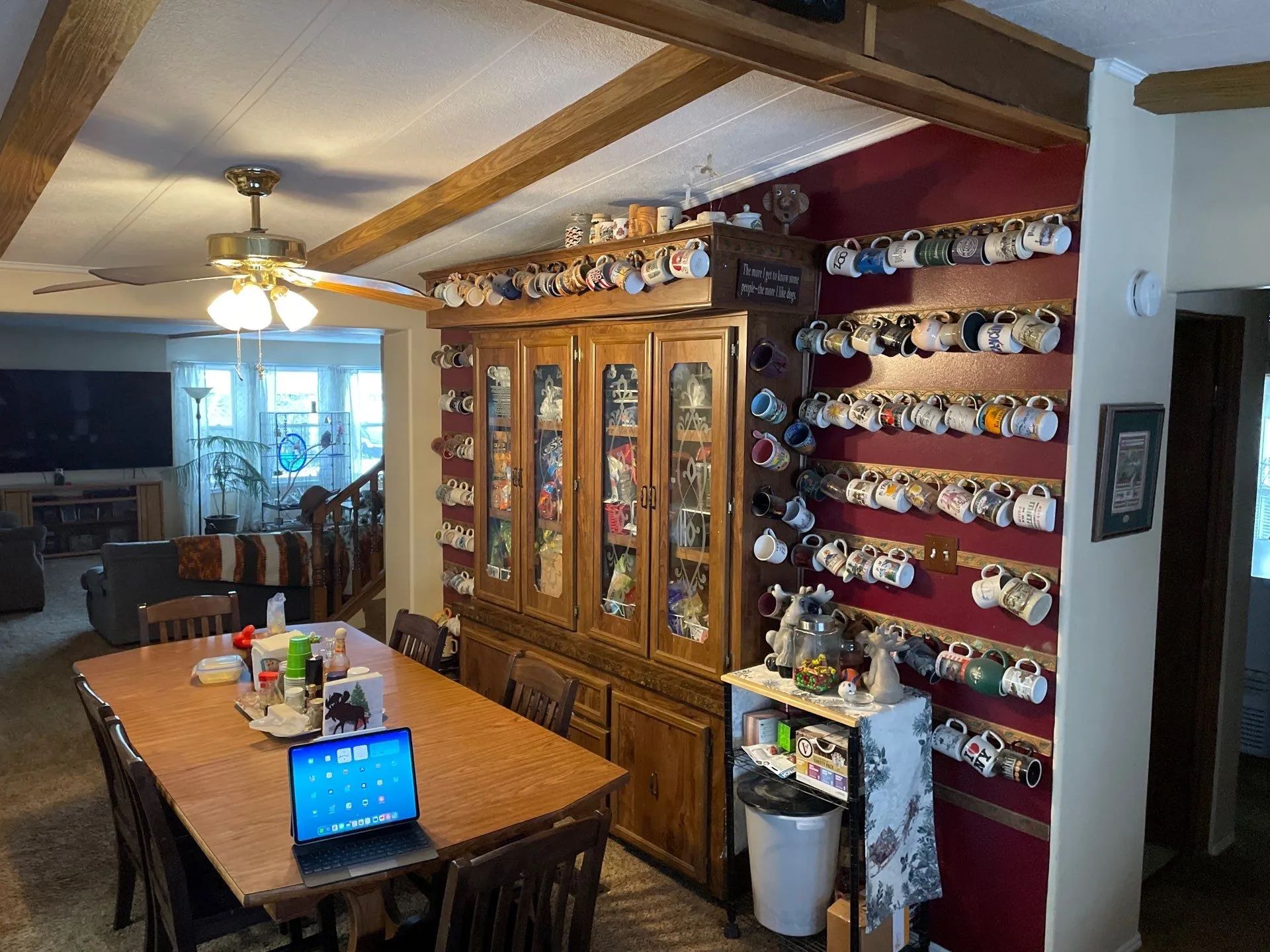Dining room with wooden table, laptop, and glass-front cabinet lined with colorful mugs and plates.