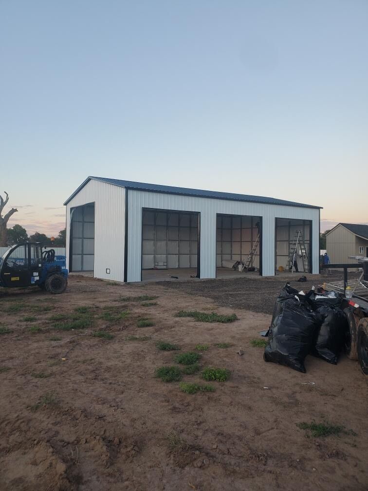 Three-bay white and black metal garage under construction, in a dirt lot, with a UTV parked nearby.