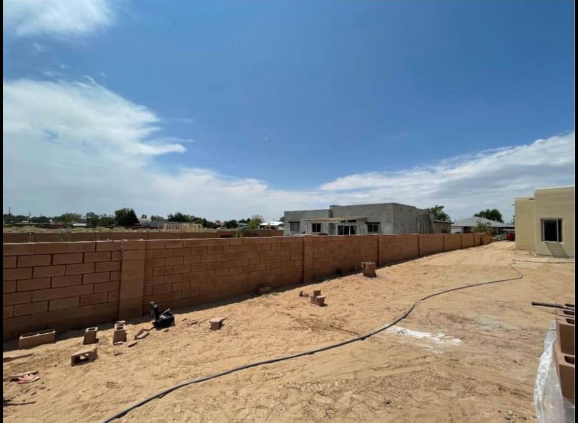A brick wall surrounds a dirt field with a house in the background