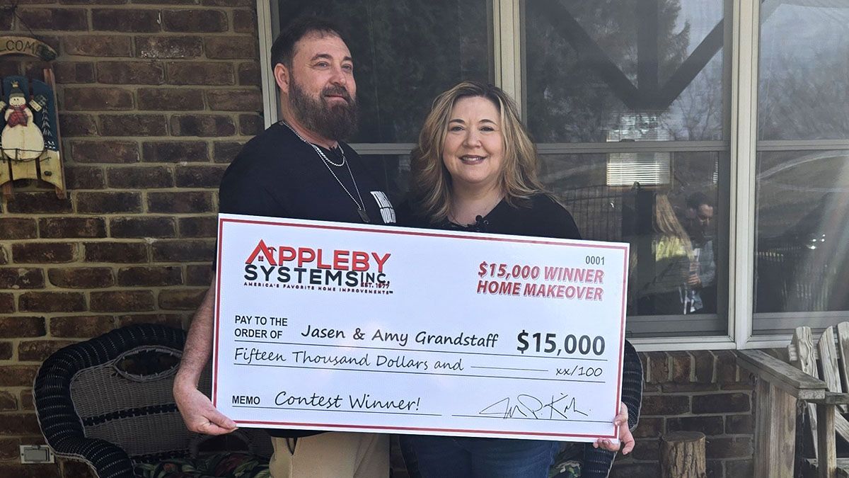 Two people holding a large $15,000 check on a porch, smiling at the camera