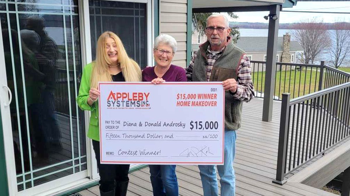 Three people hold a large check outside a building on a covered walkway