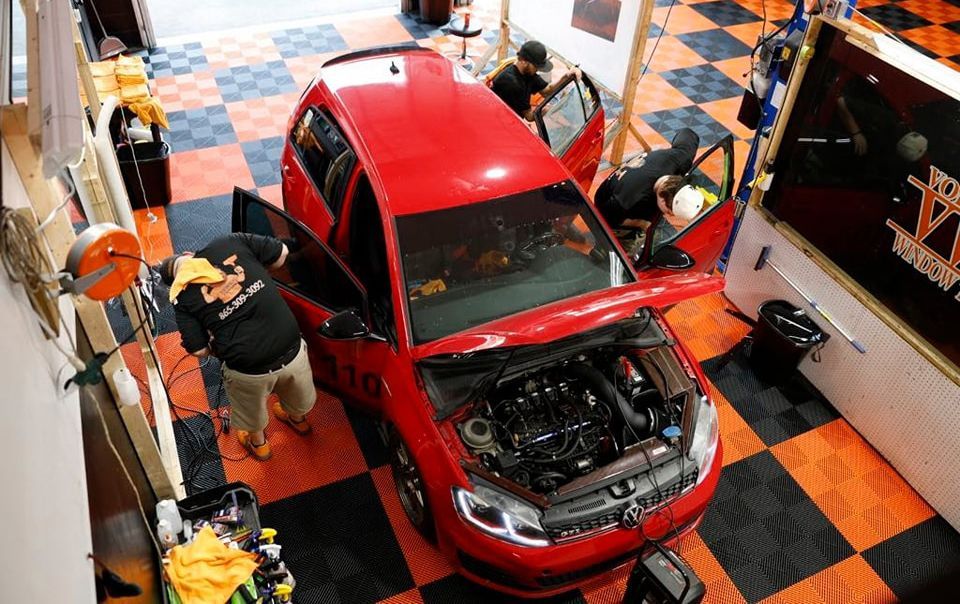 People working on a red car in a garage, with doors open. The floor is checkered black and orange.