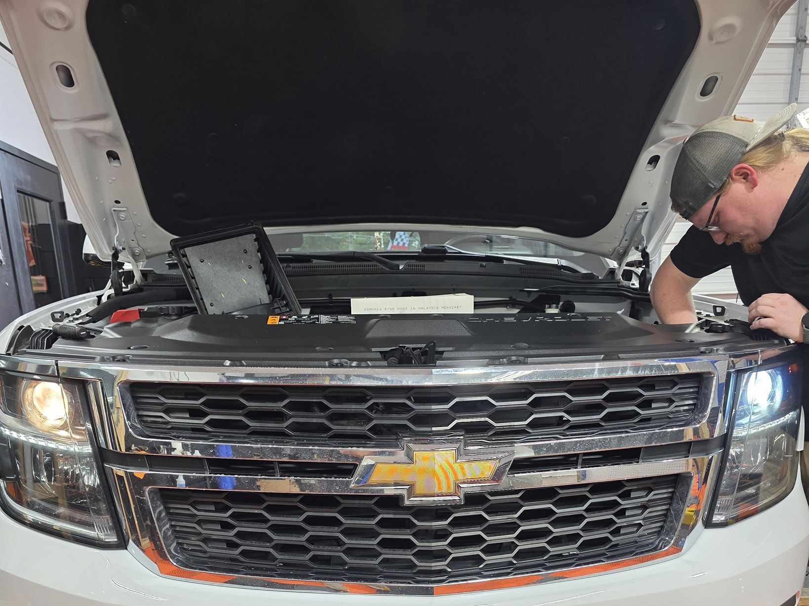 Man working on a white Chevrolet SUV engine with the hood open, in a garage.