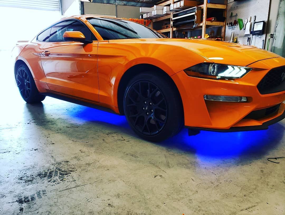 Orange Ford Mustang with blue underglow, black wheels, parked in a garage.