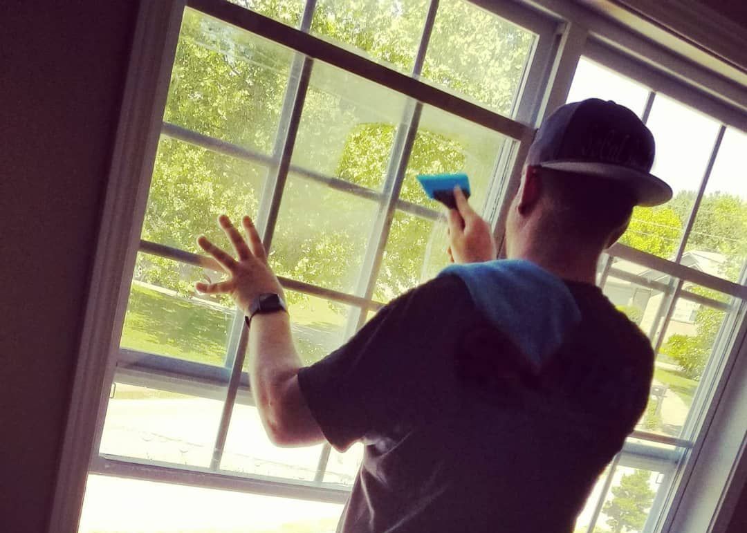 Man squeegeeing a window pane in a sunlit room; he wears a cap and watches a tree outside.