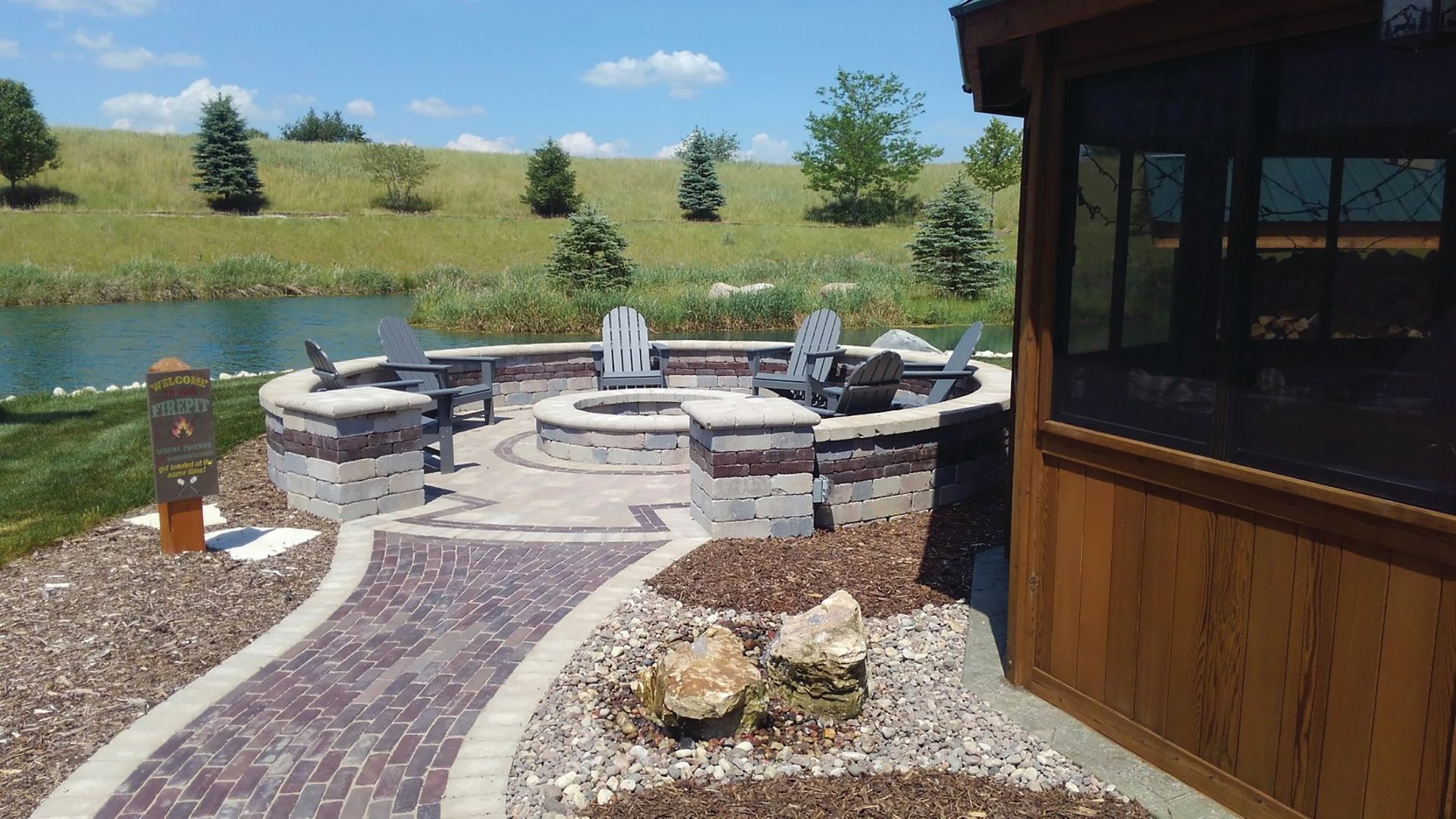 Outdoor patio with a fire pit, overlooking a pond. Brick walkway, Adirondack chairs, and a gazebo on the right.
