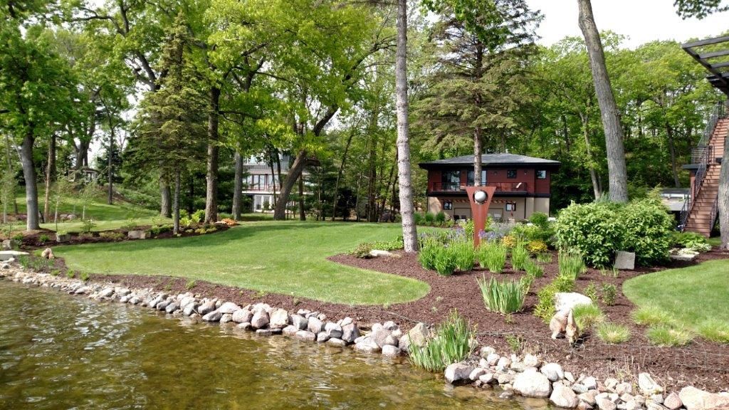 Lakeside yard with manicured lawn, dark mulch flowerbeds, stone border, trees, and a two-story brown building.