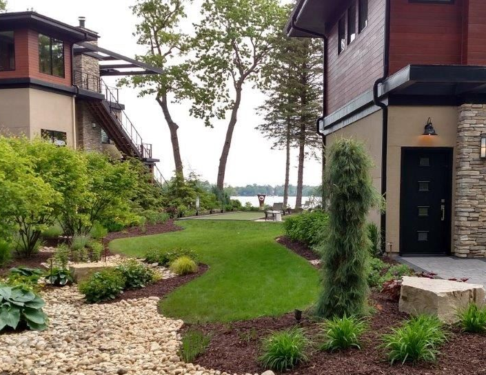 Modern home with landscaped yard leading to a lake. Green grass, brown mulch, and stone accents.