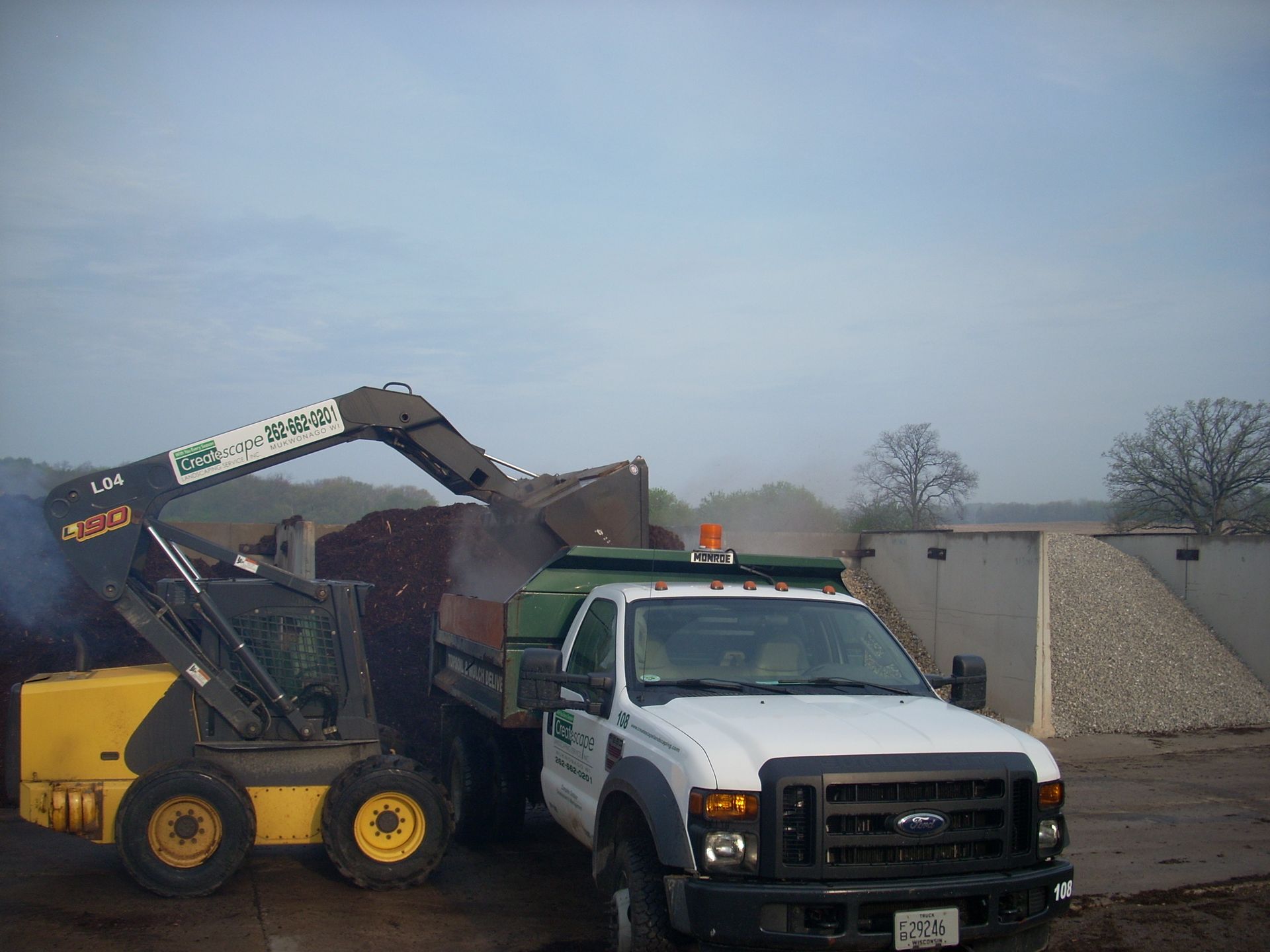 A yellow skid steer loading mulch into a white pickup truck.