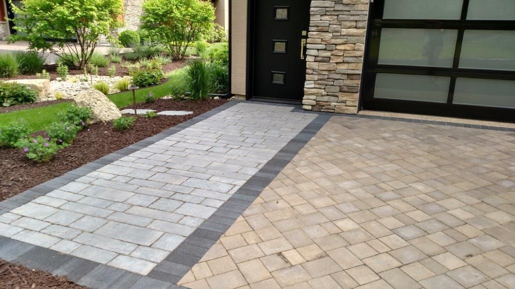 Brick walkway leading to a black door, with a tan driveway and landscaped yard.