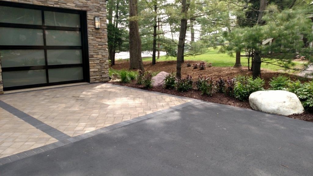 Driveway with pavers, black asphalt, and landscaped bed with large boulder next to modern garage.