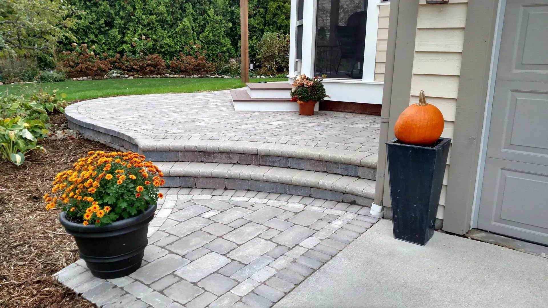 Brick patio with steps, potted mums, pumpkin, and doorway.