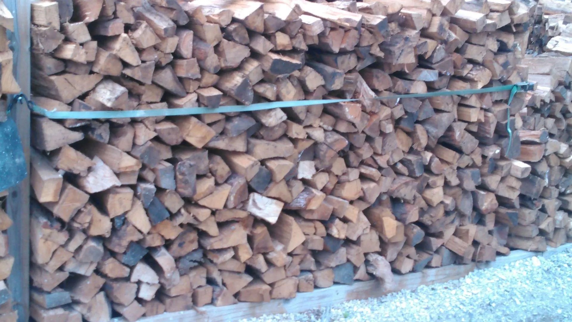 Pile of stacked firewood, secured with a green strap, outdoors.