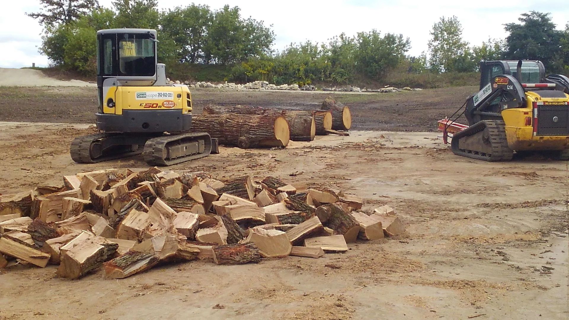 Construction site with excavator and skid steer moving logs and firewood.