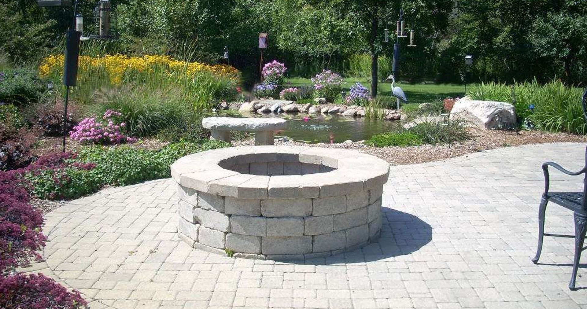 Stone fire pit on a brick patio with a pond and flowers in the background.