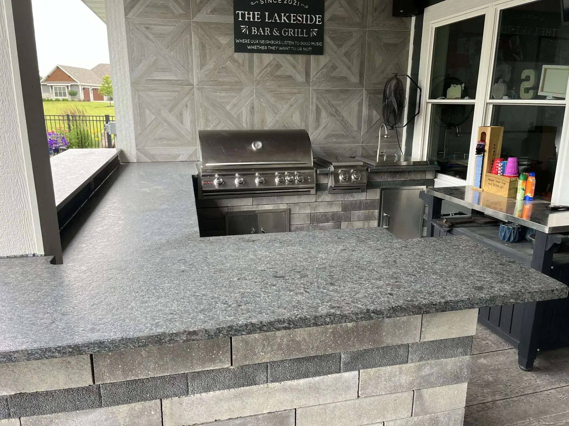 Outdoor kitchen with a built-in grill, gray countertop, and brick facade under a covered patio.