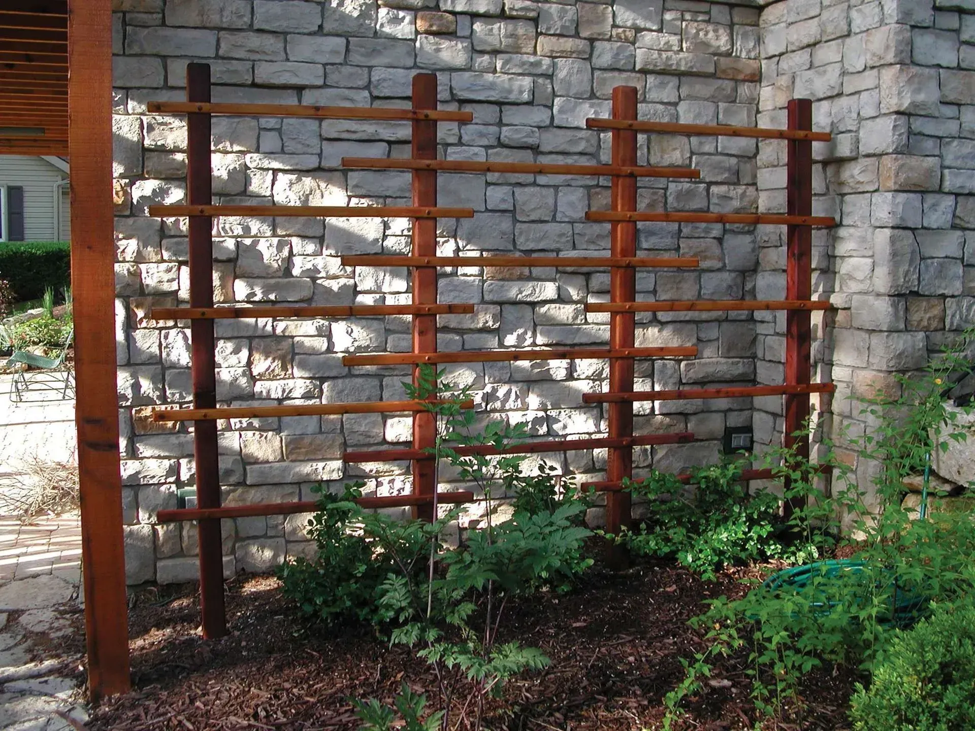 Wooden trellis against a stone wall, supporting climbing plants in a garden bed.