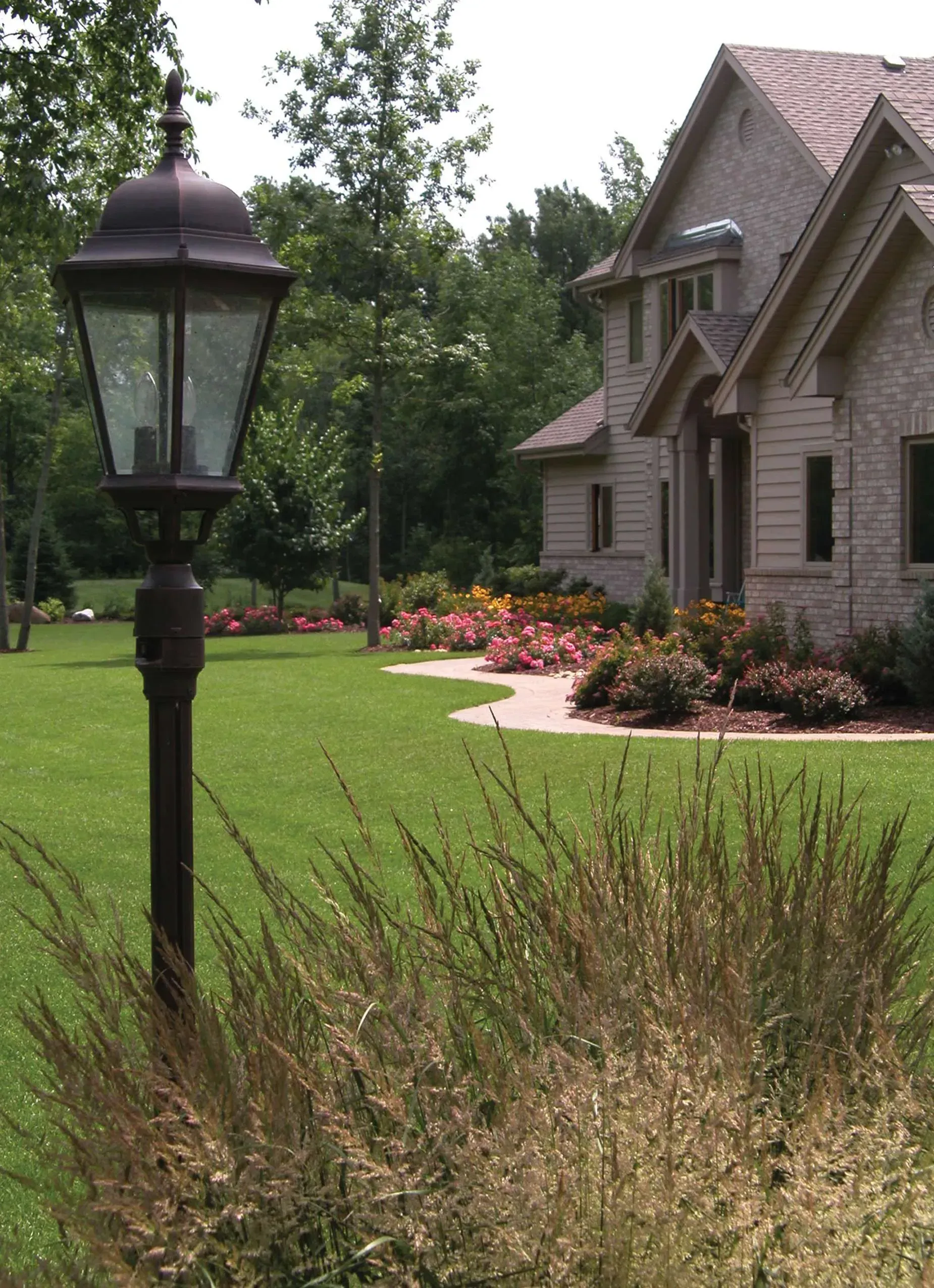 Lamp post in front of a brick house with a green lawn and colorful flower beds.
