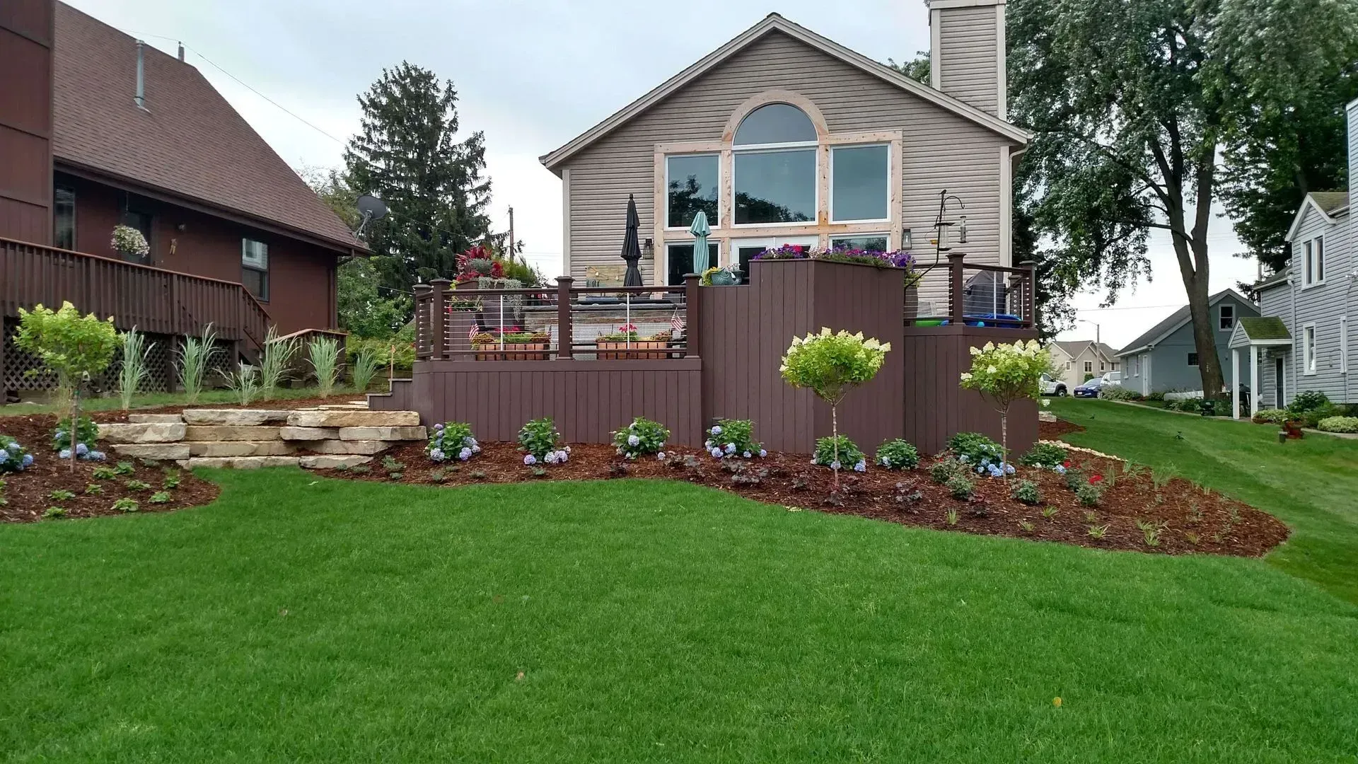 A backyard with a tiered deck, lush green lawn, and beige house. Landscaping features trees and flowers.