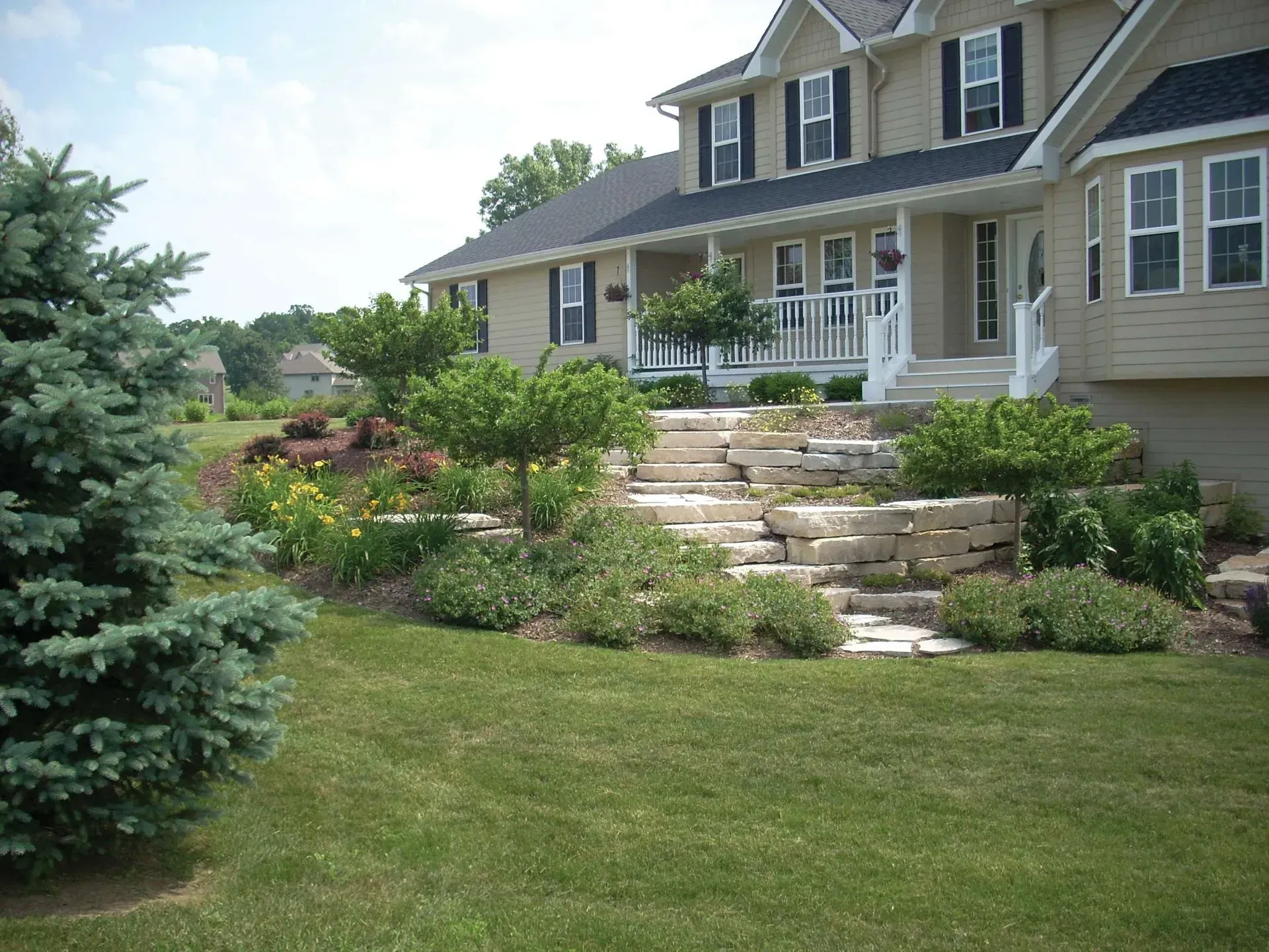 Beige house with stacked stone steps leading to a porch; landscaped yard with a blue spruce and green grass.