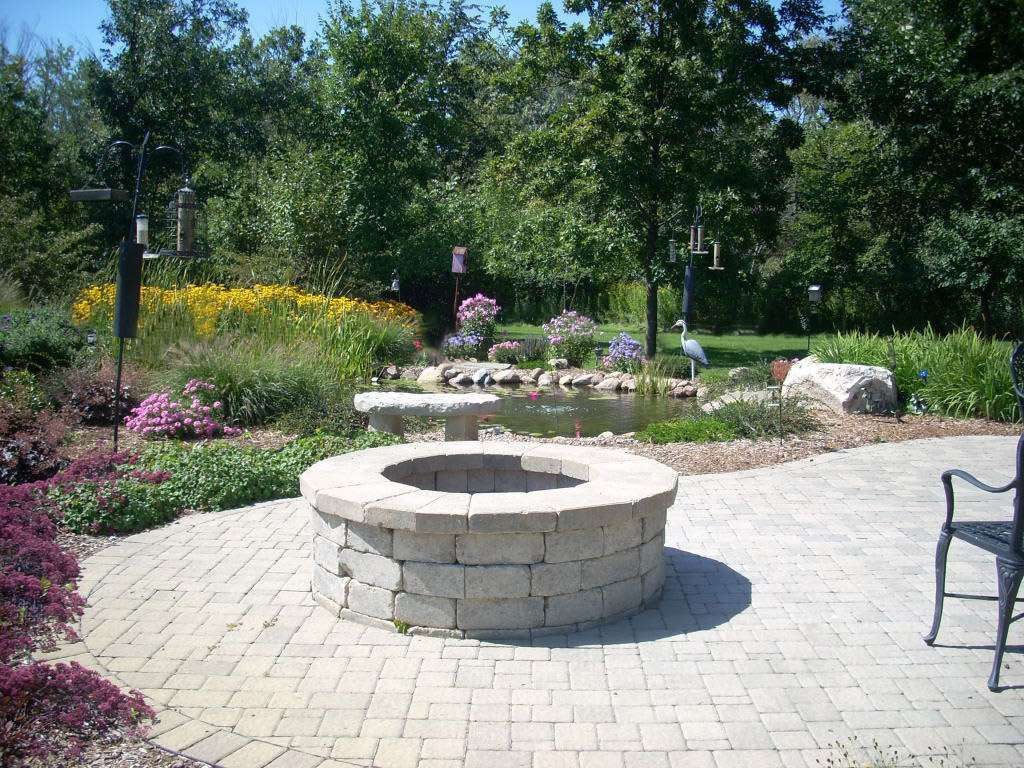Brick fire pit on a patio, overlooking a pond with flowers and trees. Sunny day.