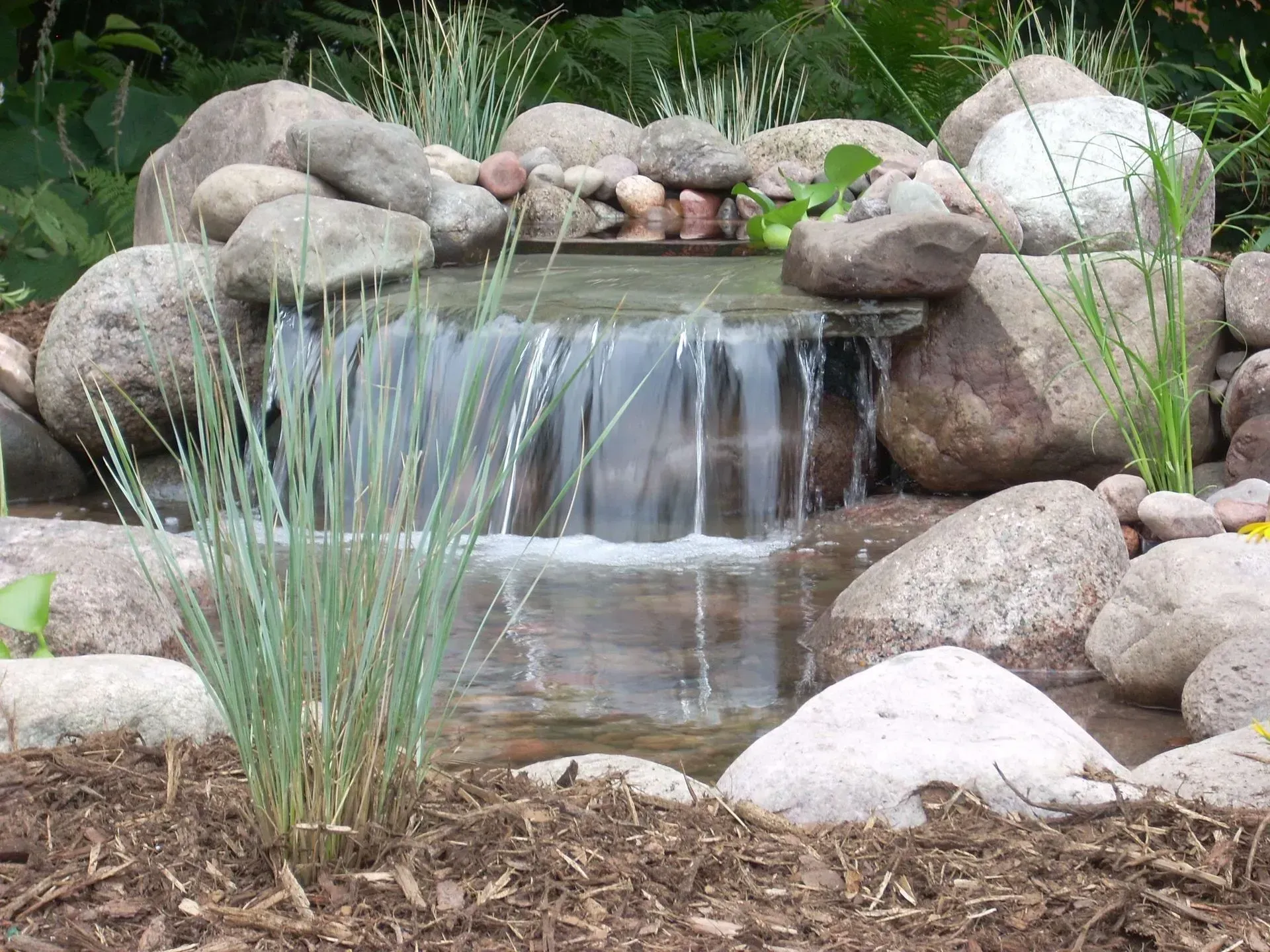Small waterfall cascading over rocks into a pond, surrounded by stones and plants.