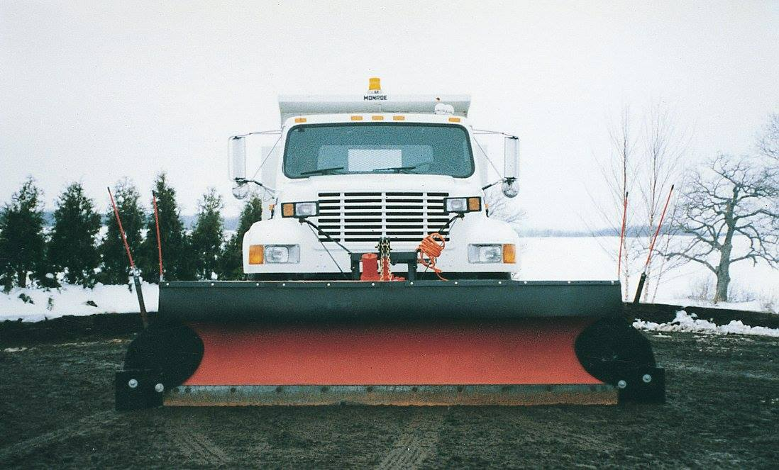 Front view of a white snowplow truck with a red blade, clearing a snowy road.