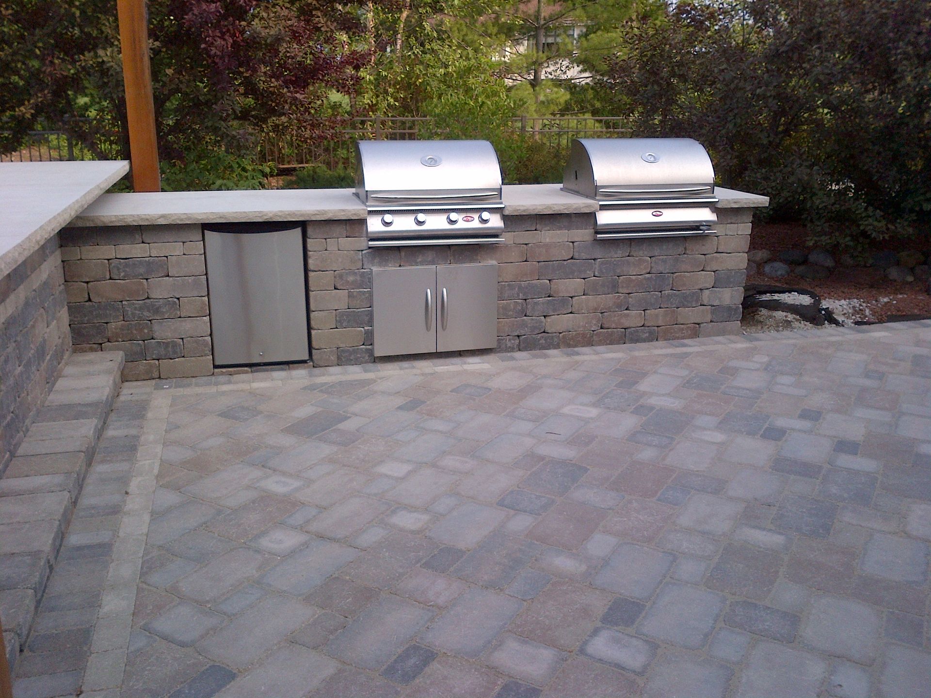 Outdoor stone kitchen with two grills, refrigerator, and cabinet on a paved patio.