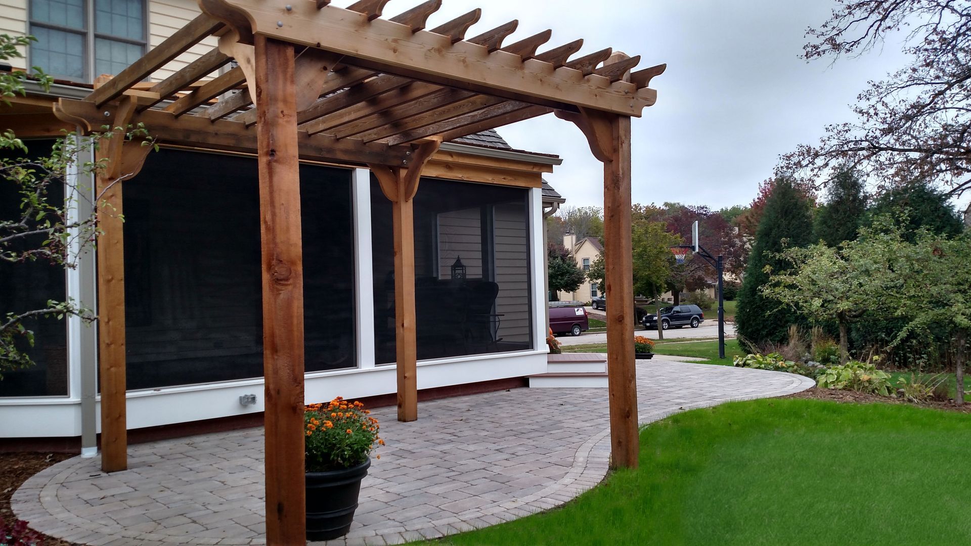 Wooden pergola over a brick patio next to a house with screening, grass, and landscaping.