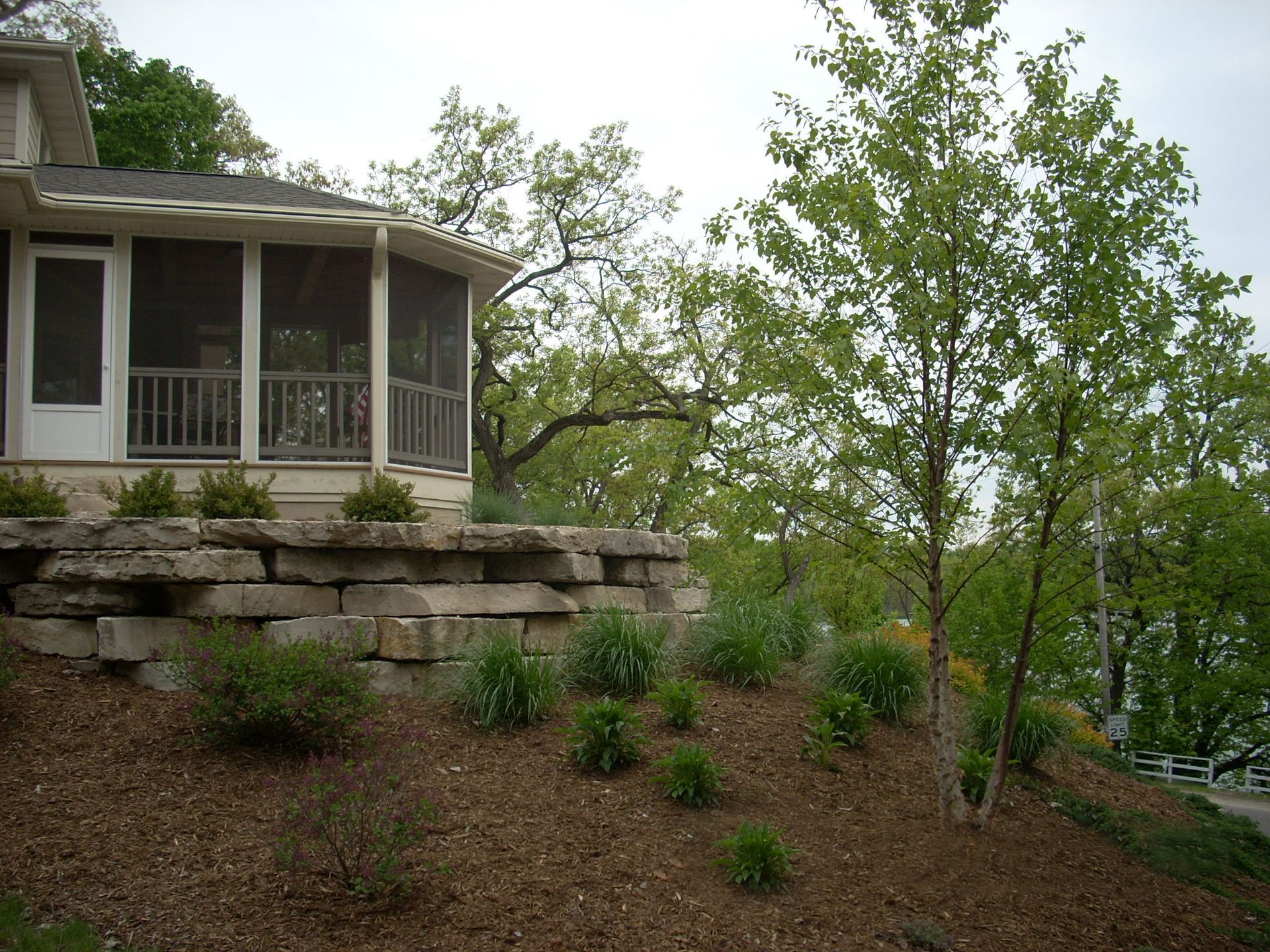 Screened porch, stone retaining wall, and landscaped hillside with young trees.
