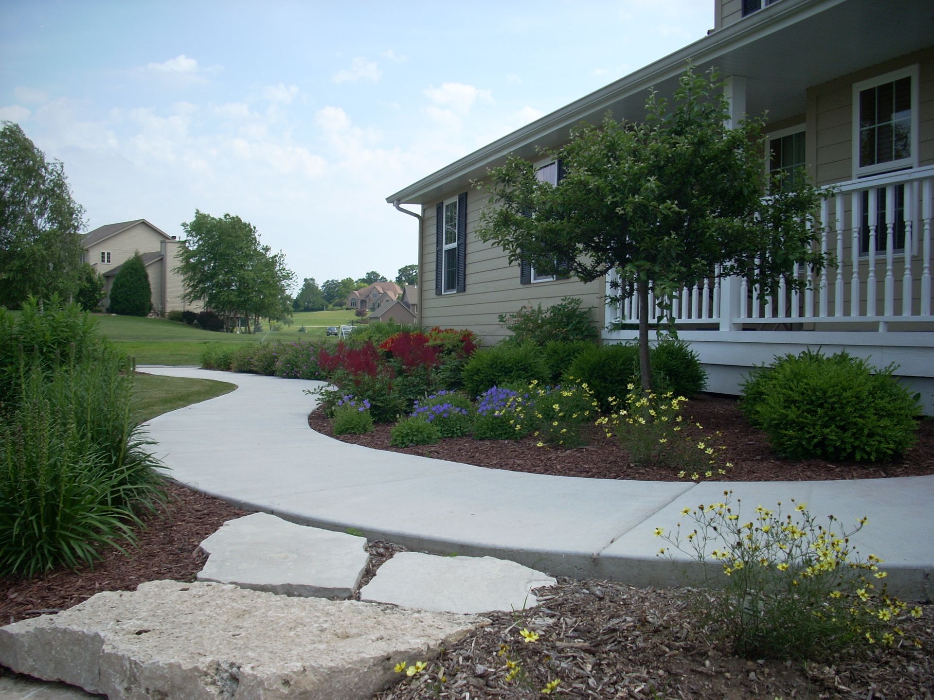 Curving sidewalk leads past a house with a porch, surrounded by landscaping, under a partly cloudy sky.