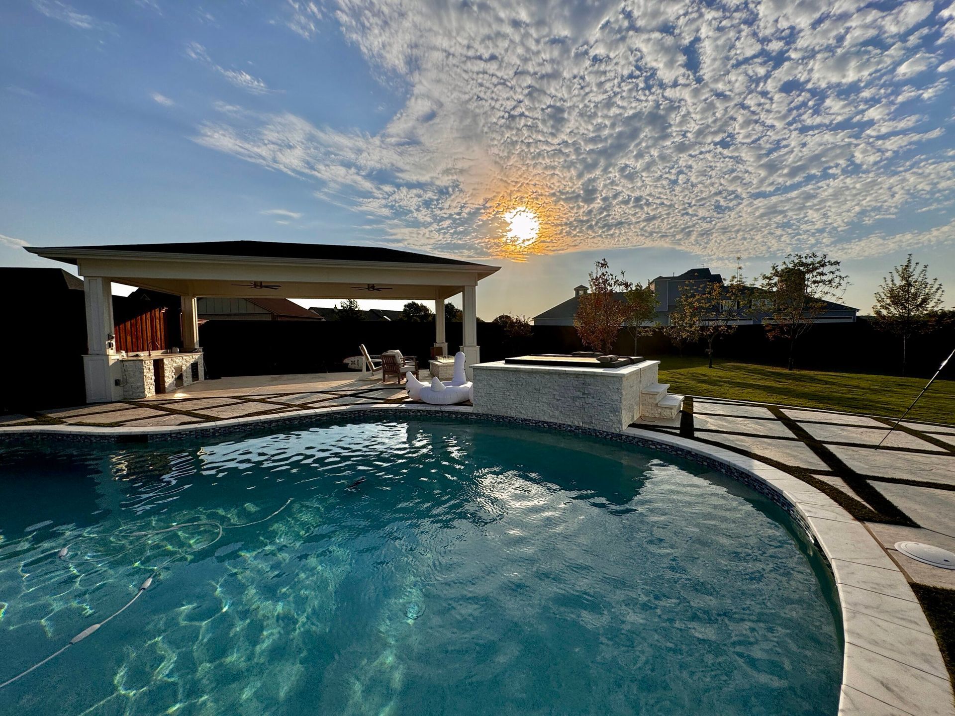 A large swimming pool with a gazebo in the background and the sun shining through the clouds.