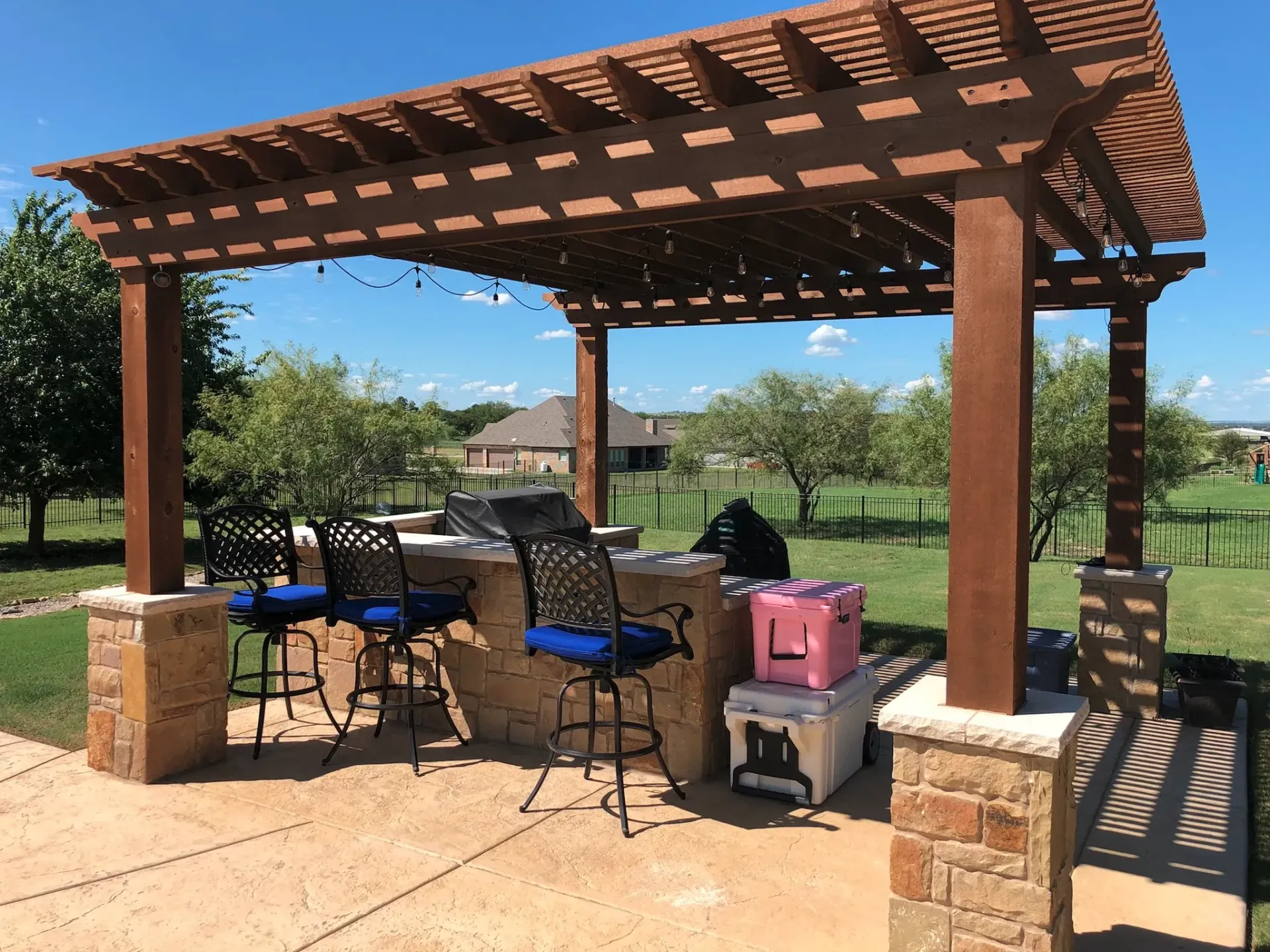 A wooden pergola with a grill and chairs underneath it