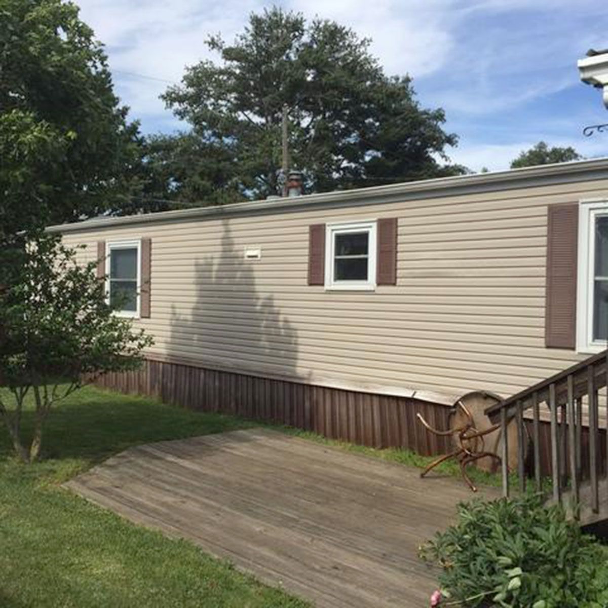 Tan mobile home with brown shutters and trim, wooden deck, green grass, and trees