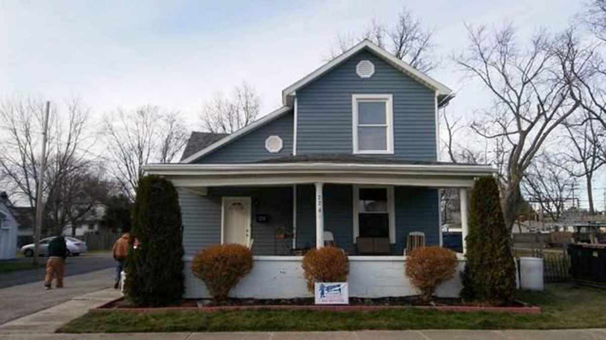 Blue two-story house with porch; two people, bare trees, and shrubs in front; overcast day
