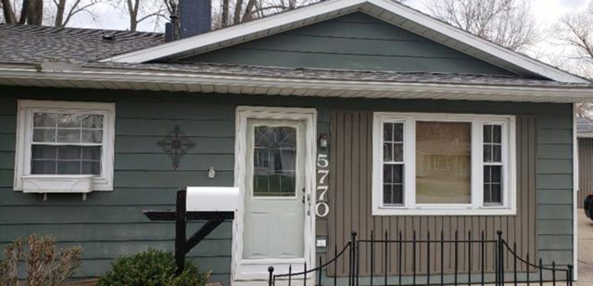Green and brown house with a white door, windows, and mailbox; black wrought iron fence
