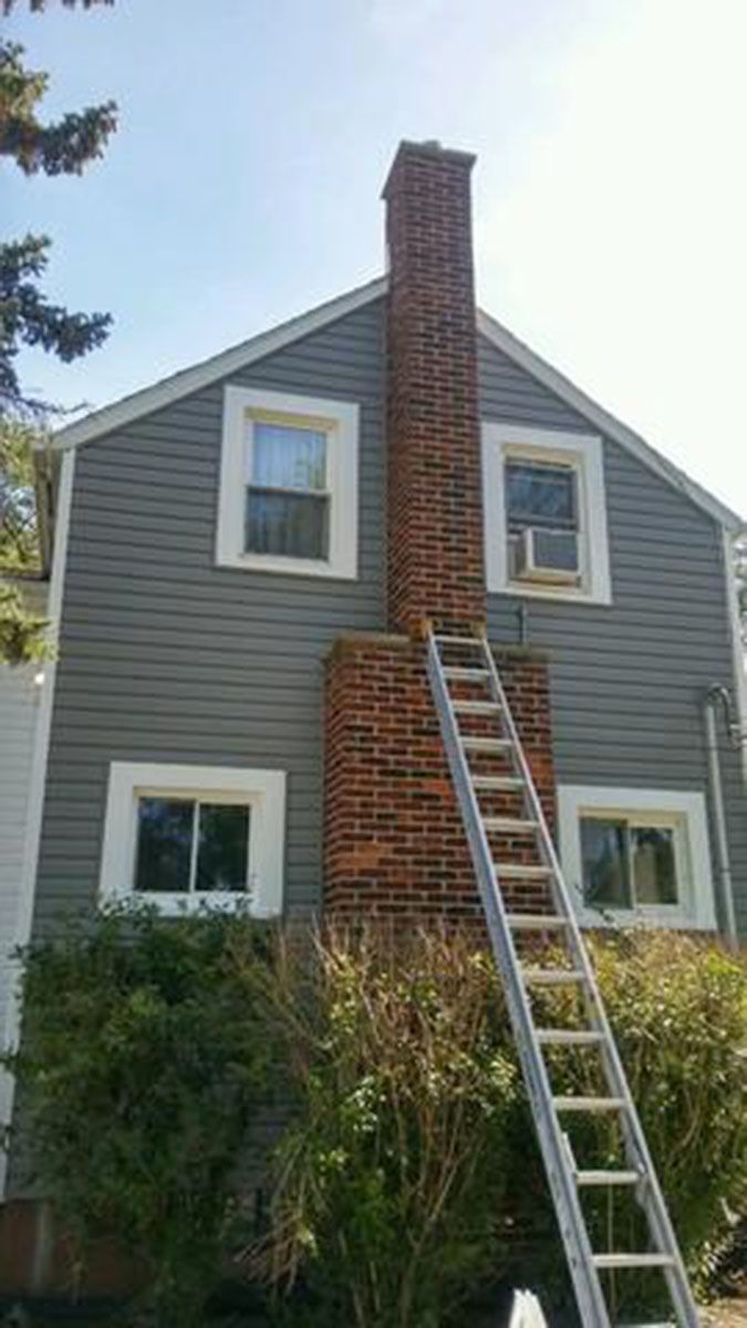 Ladder leaning against brick chimney on the side of a gray-sided house with white window frames