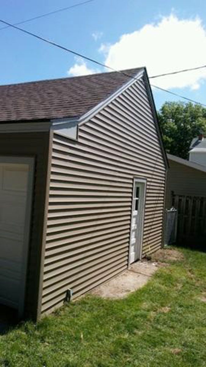 Tan-sided garage with brown shingle roof, white door, and grassy yard under blue sky