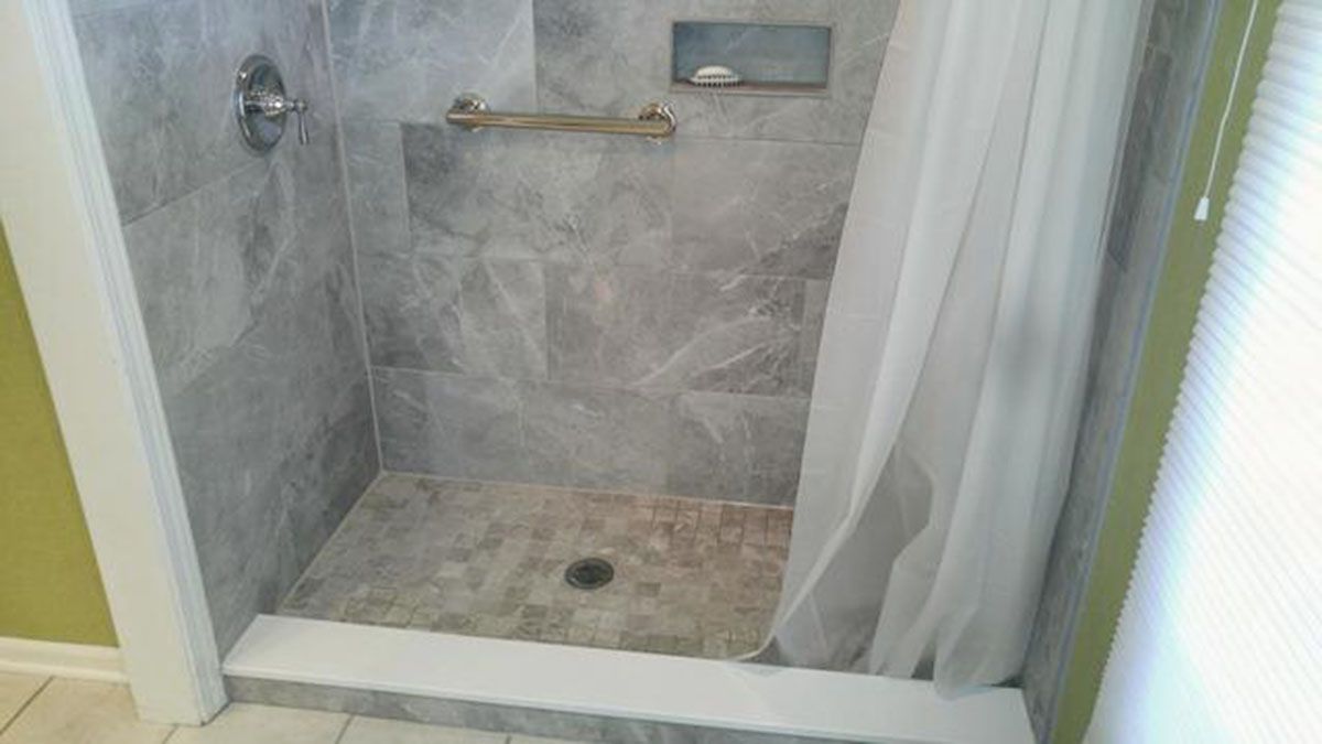 Gray tiled shower with a white curtain, grab bar, and recessed shelf