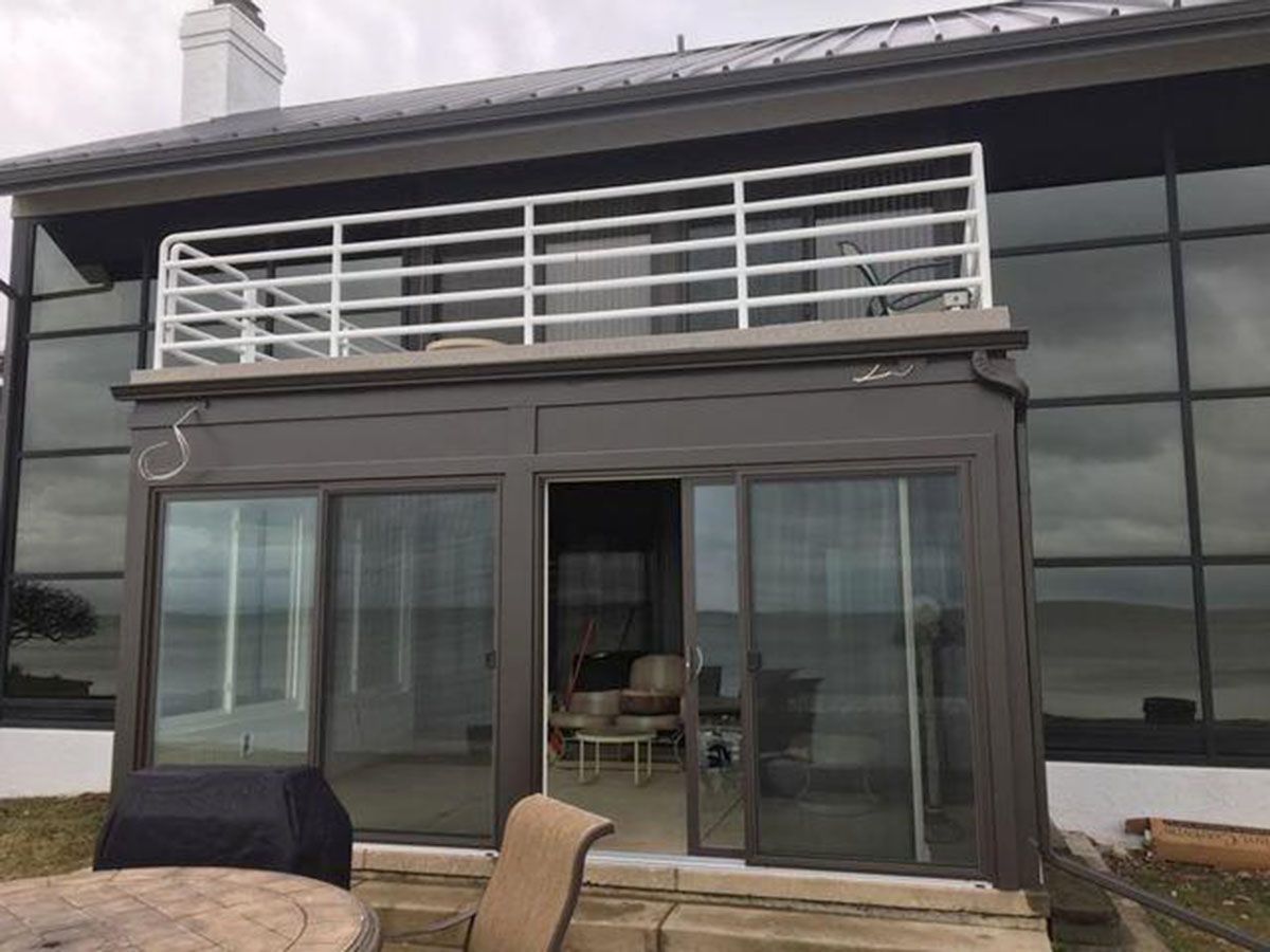 Dark-roofed house with second-story white railing and gray-walled sunroom with sliding doors and outdoor patio