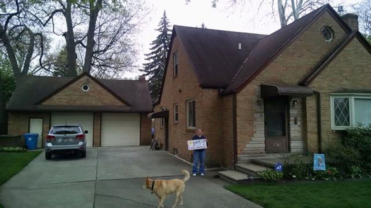 A person holding a sign stands near the front door of a brick house with a detached garage