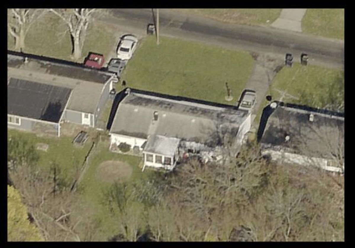 Aerial view of several houses with cars parked on a grassy lot, trees, and a road