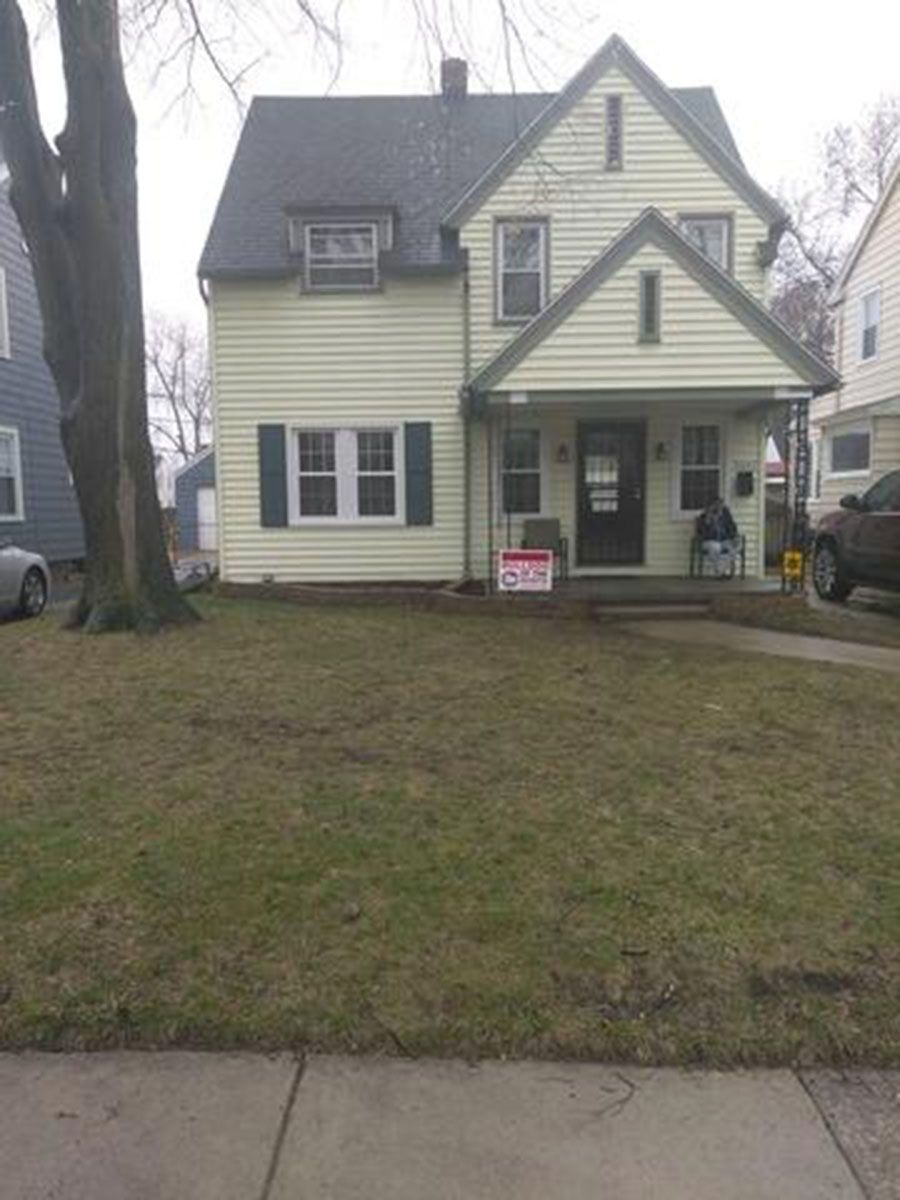 Two-story light yellow house with front porch, green shutters, and a bare tree in the front yard