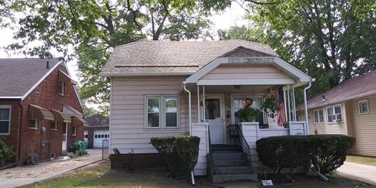Small beige house with porch, shrubs, and steps. Brick house on the left, trees in the background