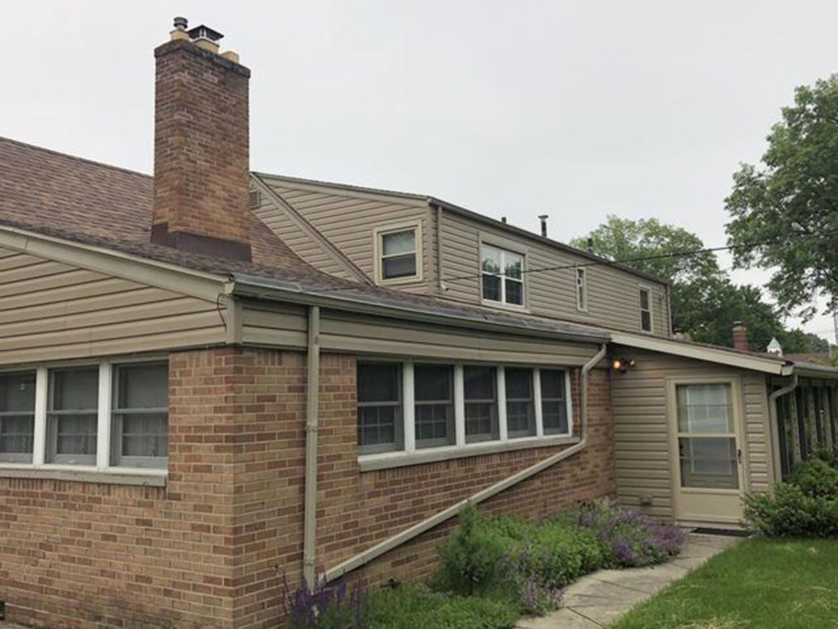 Brick house with brown roof, chimney, siding, and windows; cloudy sky