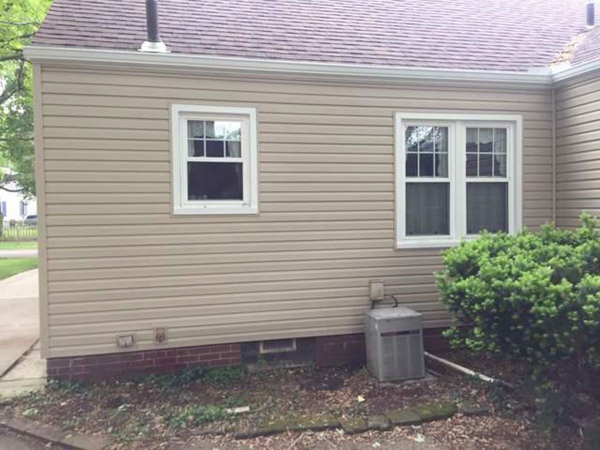 Tan siding on a house with white-framed windows, a small bush, and an AC unit